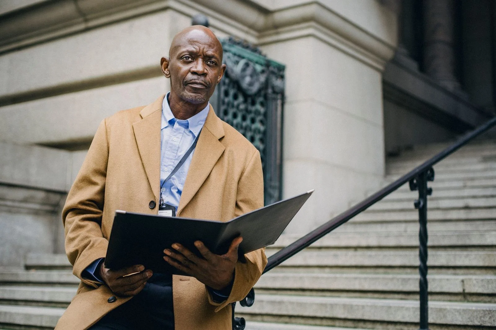 A pensive middle-aged Black man sits on a stair railing on a city street, holding an open folder and looking away.