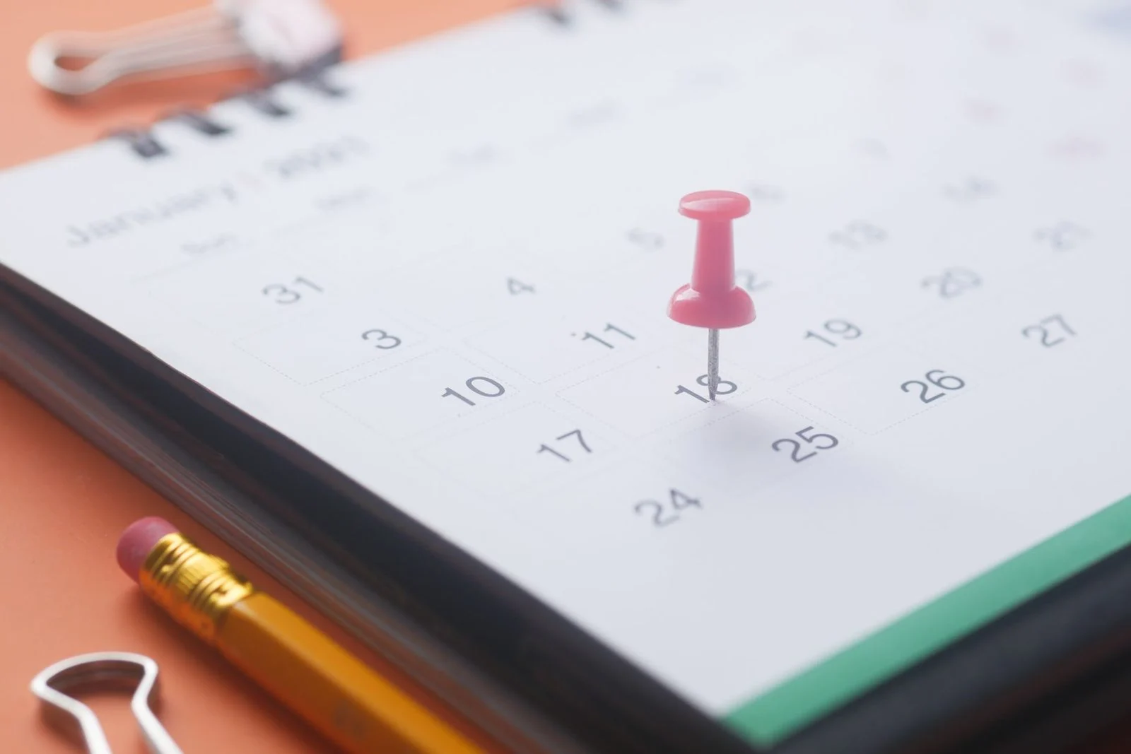 A pink push pin, pencil, and binder clip rest on a calendar page against an orange background.