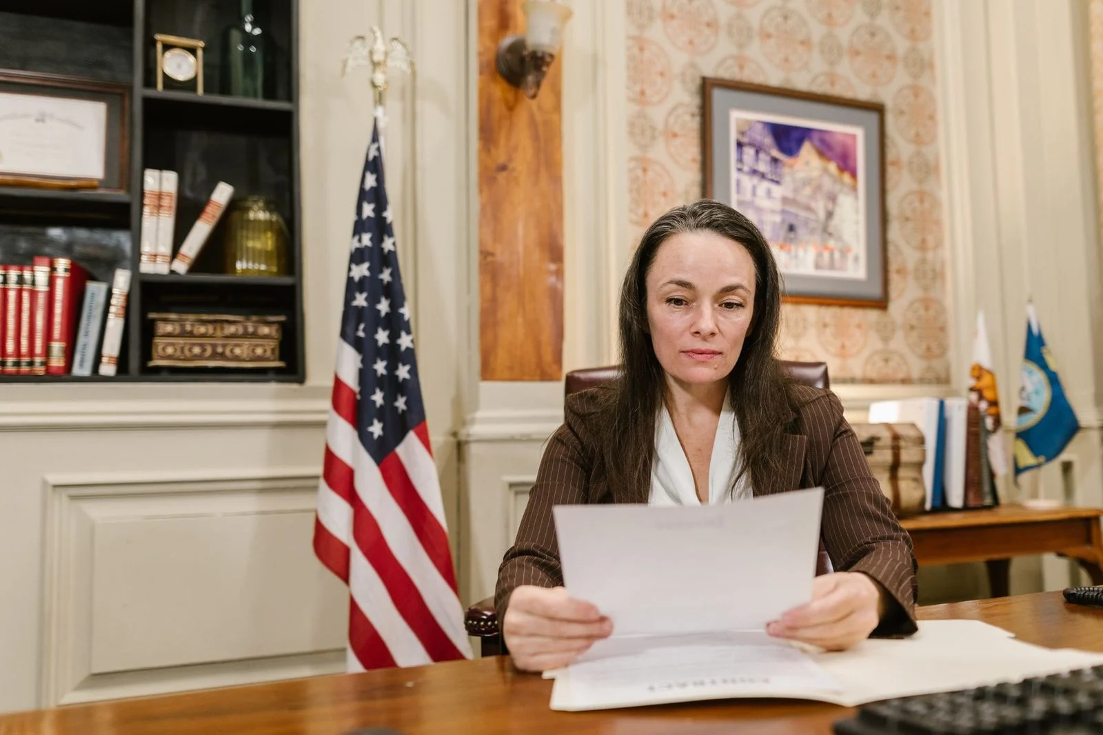 A professional woman reviews legal documents at a desk in an office with flags and bookshelves.