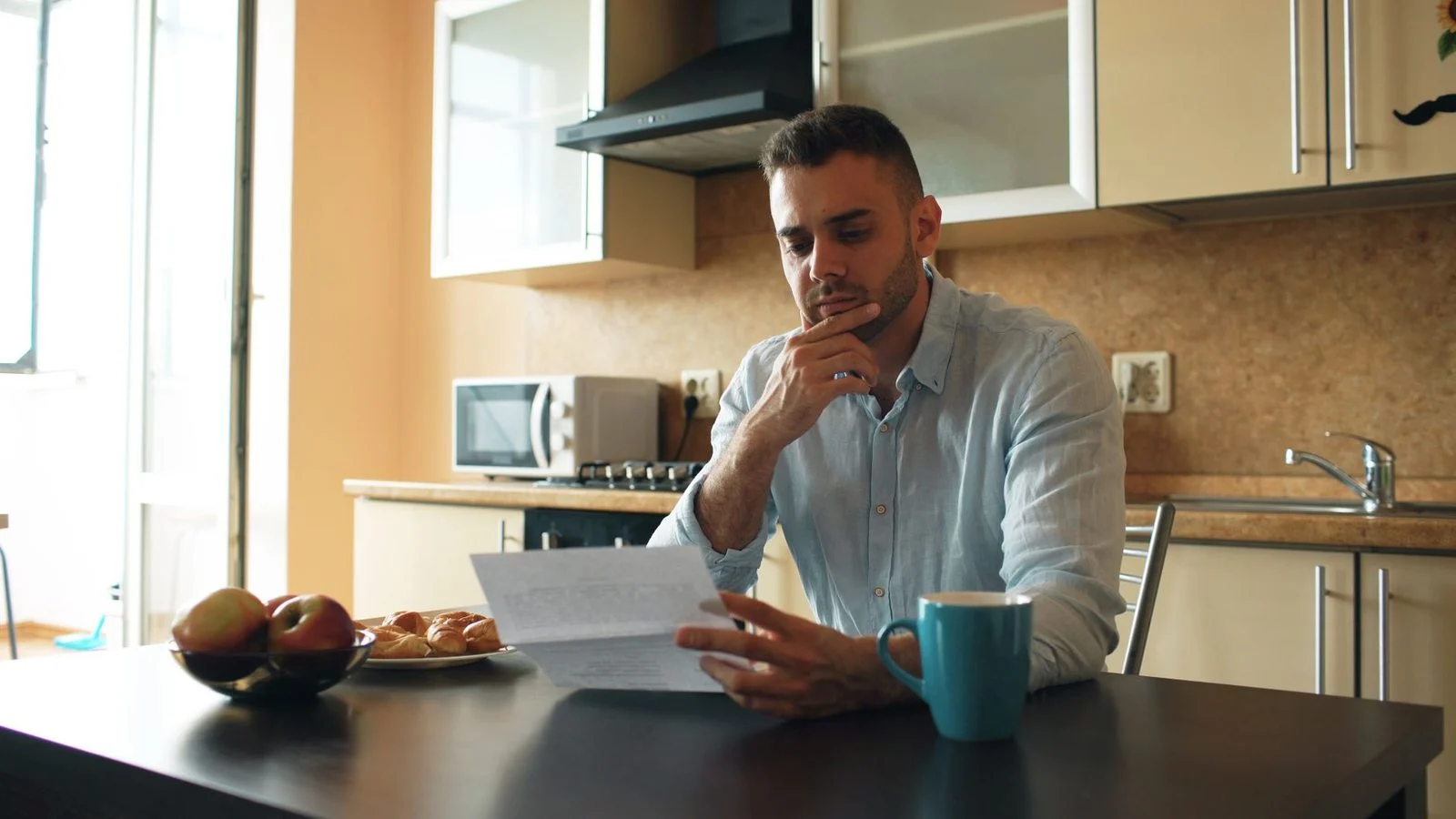 A thoughtful man reads a letter at a kitchen table in warm sunlight.