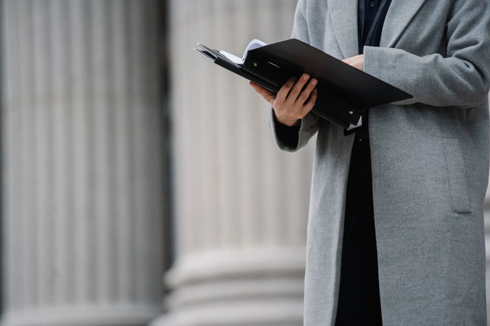 A woman in a gray coat reads a document from an open folder while standing on an autumn street.