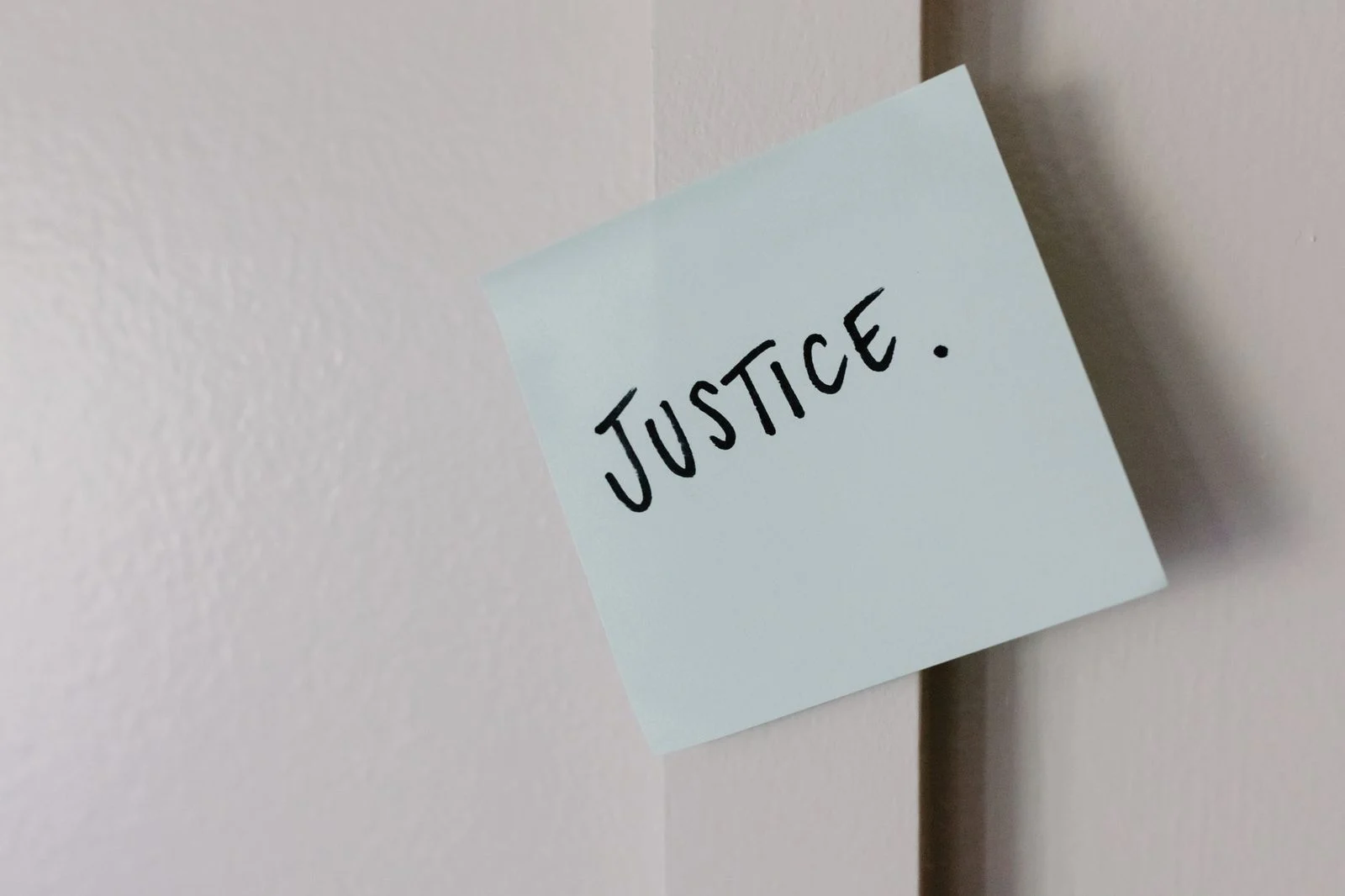 A yellow sticky note with the handwritten word "Justice" pinned to a plain white wall.