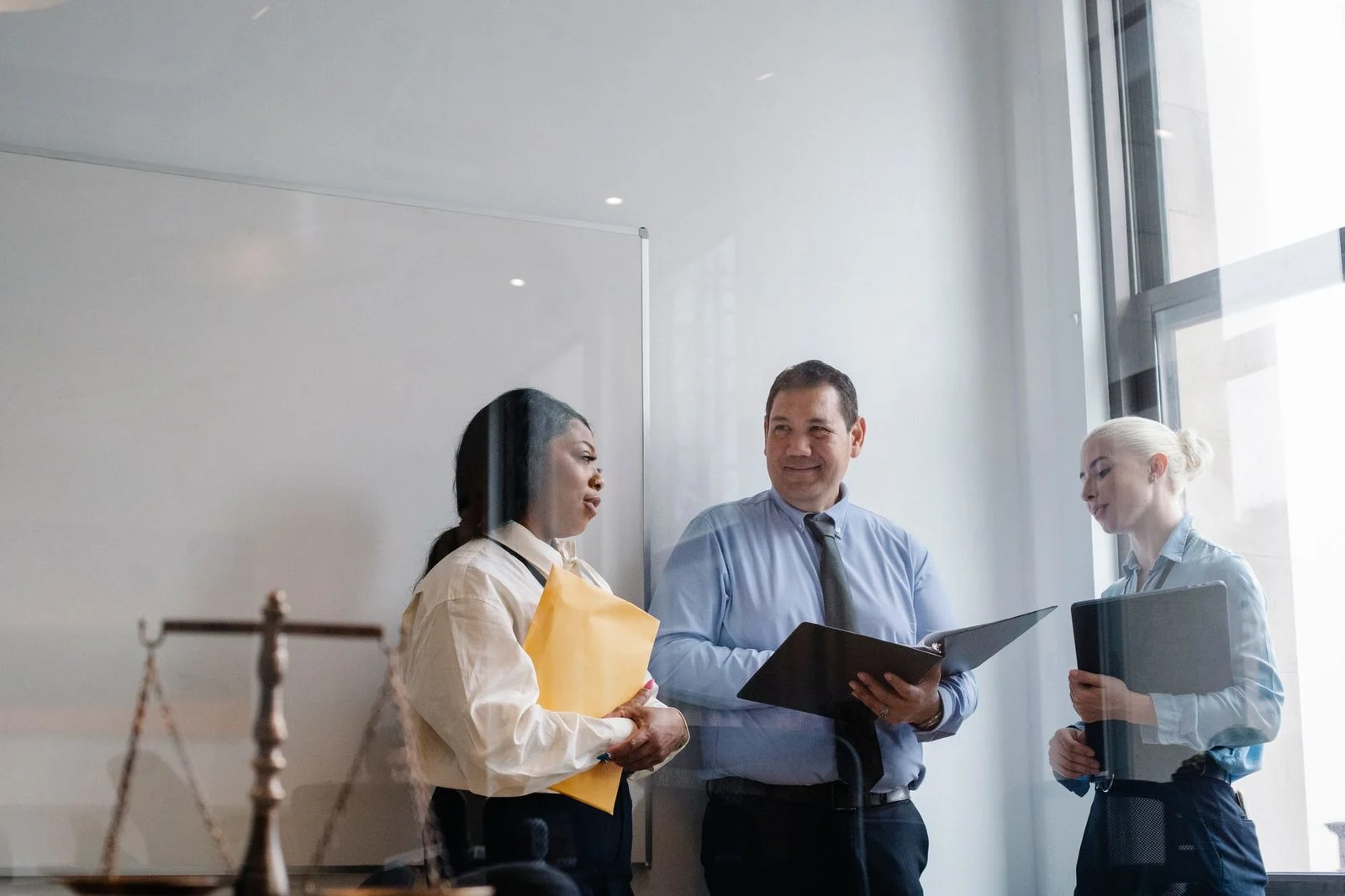 Four professionals in formal clothes stand together in an office, reviewing documents and discussing a case, photographed through glass.