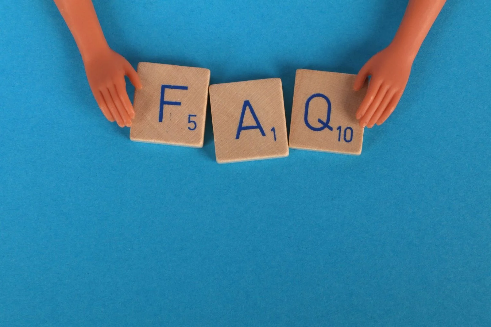 Letras de Scrabble que deletrean "FAQ" junto a pequeñas manos de juguete sobre un fondo azul.