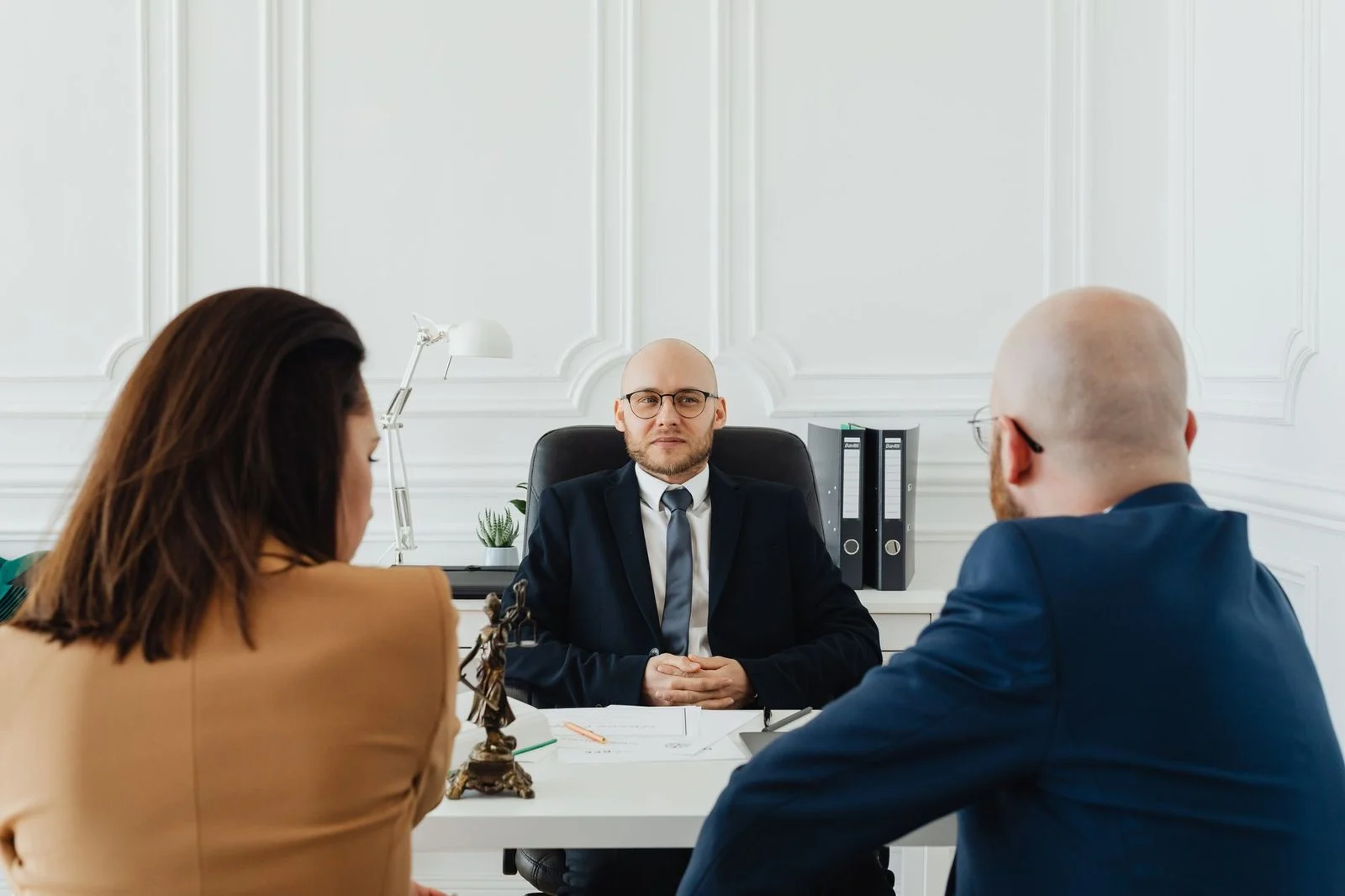 Two people sit across a desk from a lawyer in a formal office during a legal consultation.