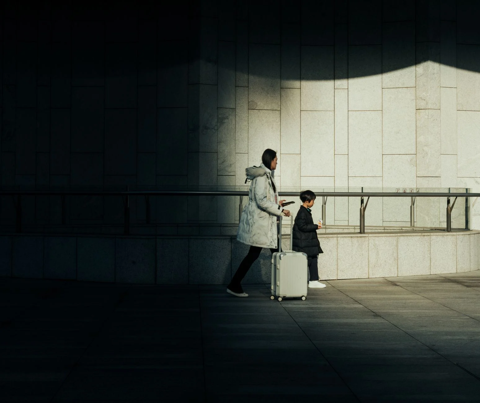 Woman and child in winter coats walking with a suitcase through a modern building.