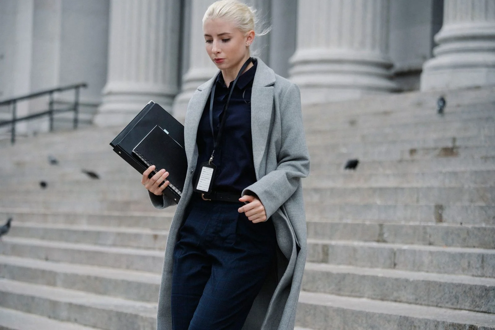 Young woman in formal clothes and a name tag carries folders down stairs beside a stone building with columns.